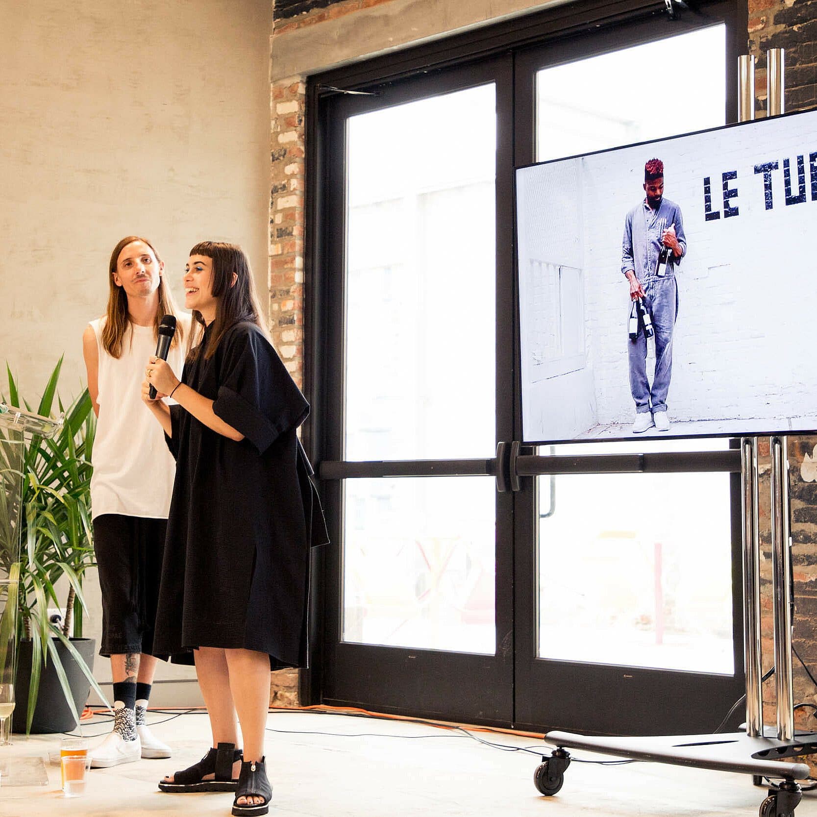 Two presenters stand in front of a screen displaying the text "LE TURTLE". One is speaking into a microphone, while the other looks on with a smile. The room has exposed brick walls and large windows, allowing natural light. A potted plant and a coffee cup are visible nearby.