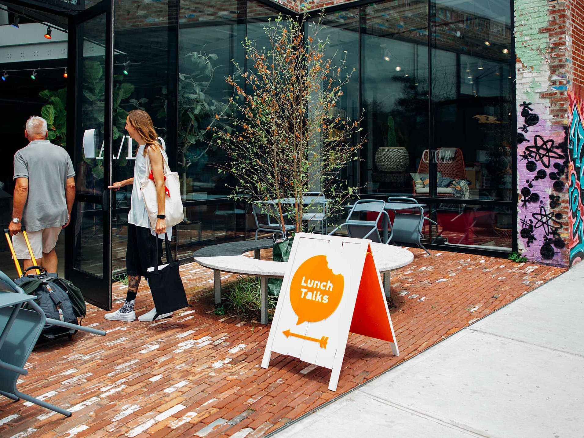 Two people are walking into a glass-fronted building with a sign that reads "Lunch Talks" on a sidewalk. The storefront is modern and has a mix of brick and glass. The scene includes outdoor seating and vibrant graffiti art on the adjacent wall.