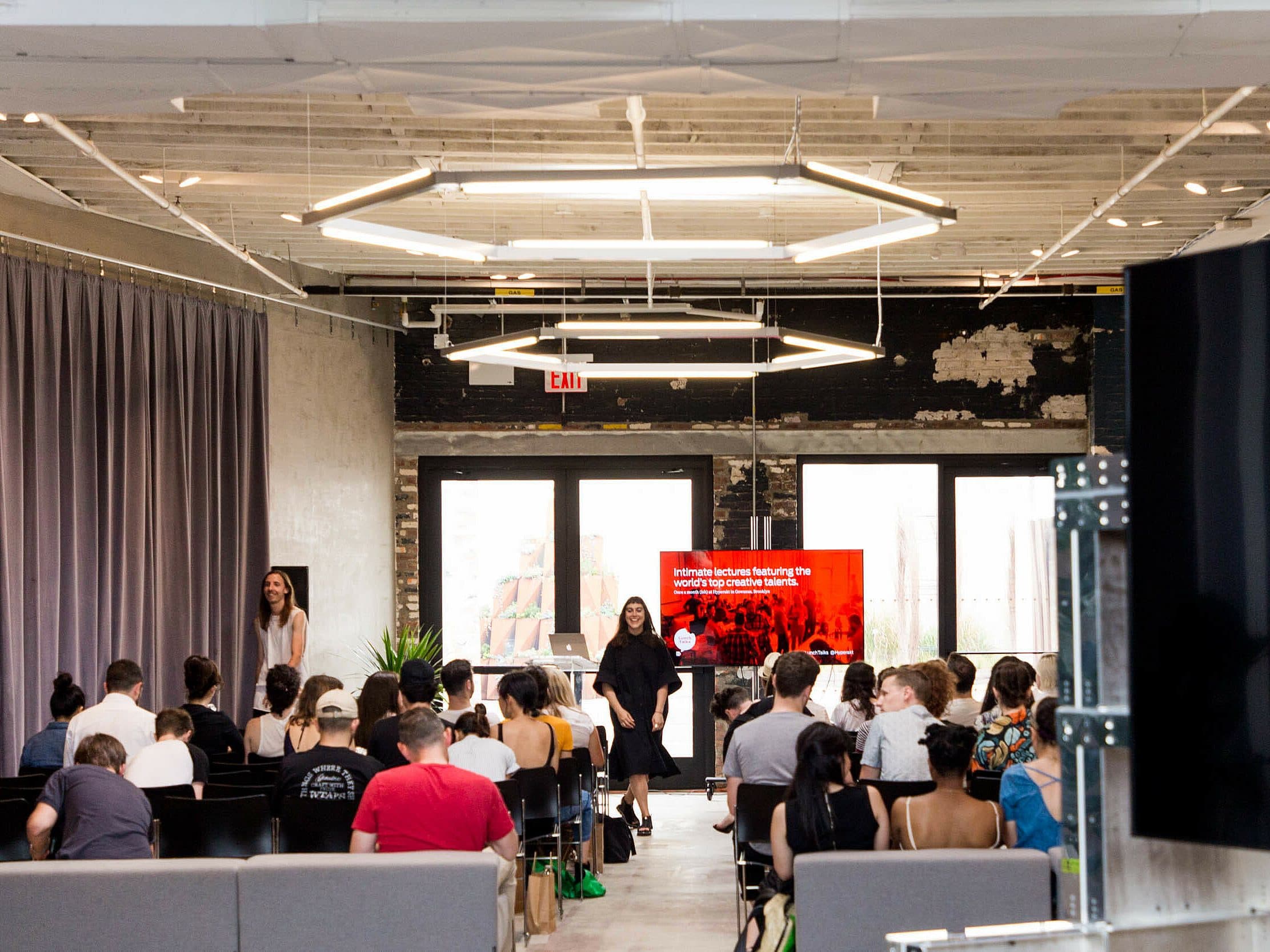 A group of people sits facing a presentation screen in a modern, industrial-style room. Two presenters stand near the screen, which displays a red background with white text. The room has large windows, exposed brick walls, and geometric light fixtures overhead.