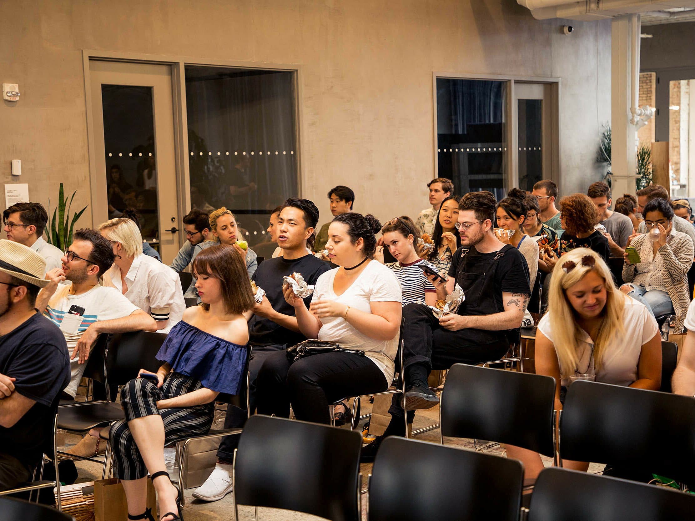 A diverse group of people sitting in rows of black chairs at an indoor event. Some are eating, chatting, or looking at their phones. The environment is casual, with a mix of attentive and relaxed demeanors. Windows and a door are visible in the background.