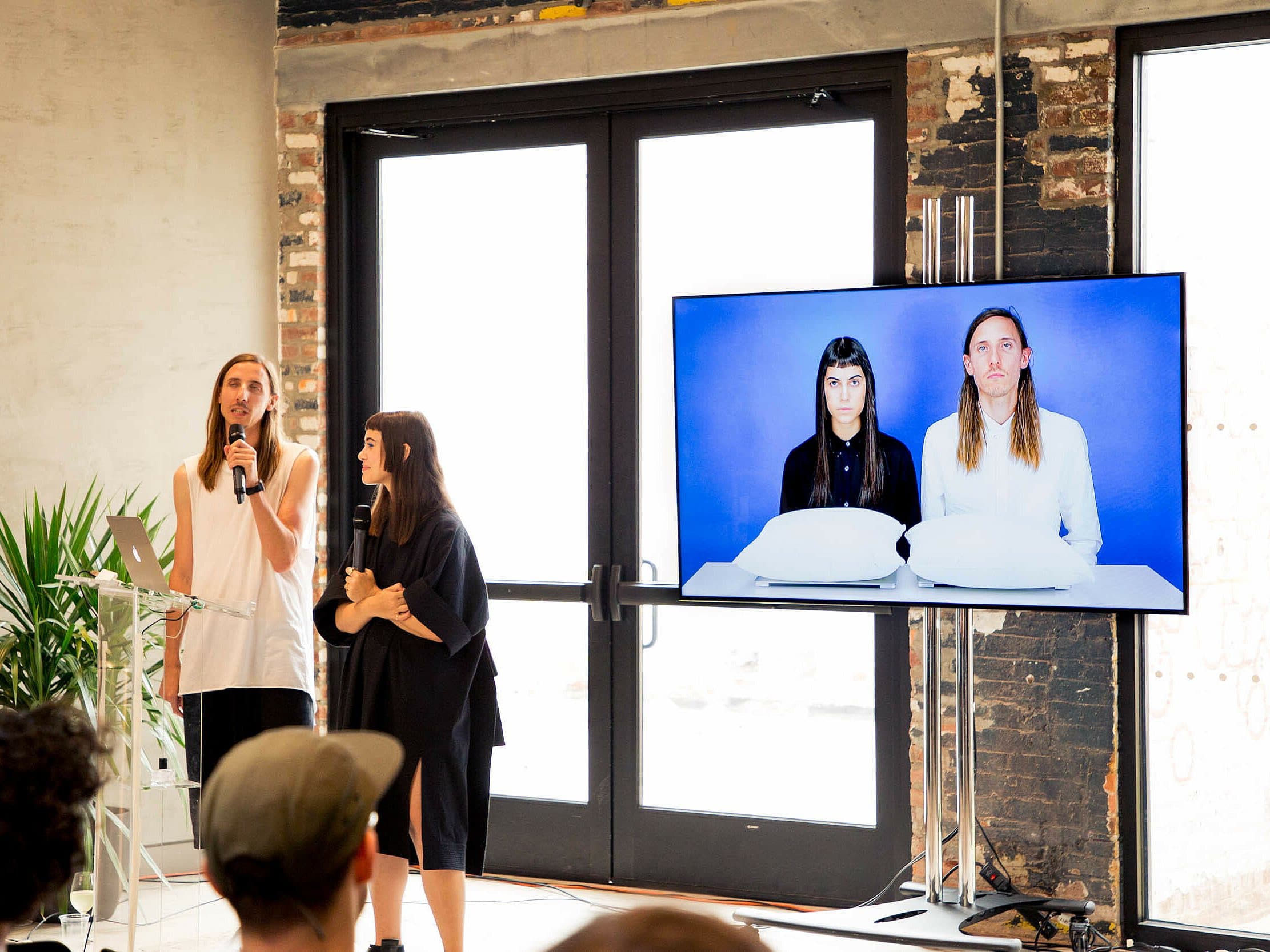 Two individuals are presenting in a modern, well-lit room with large windows. One person speaks into a microphone while the other stands beside them. A screen shows a photo of the two presenters sitting at a table with white cushions. An audience is partially visible.