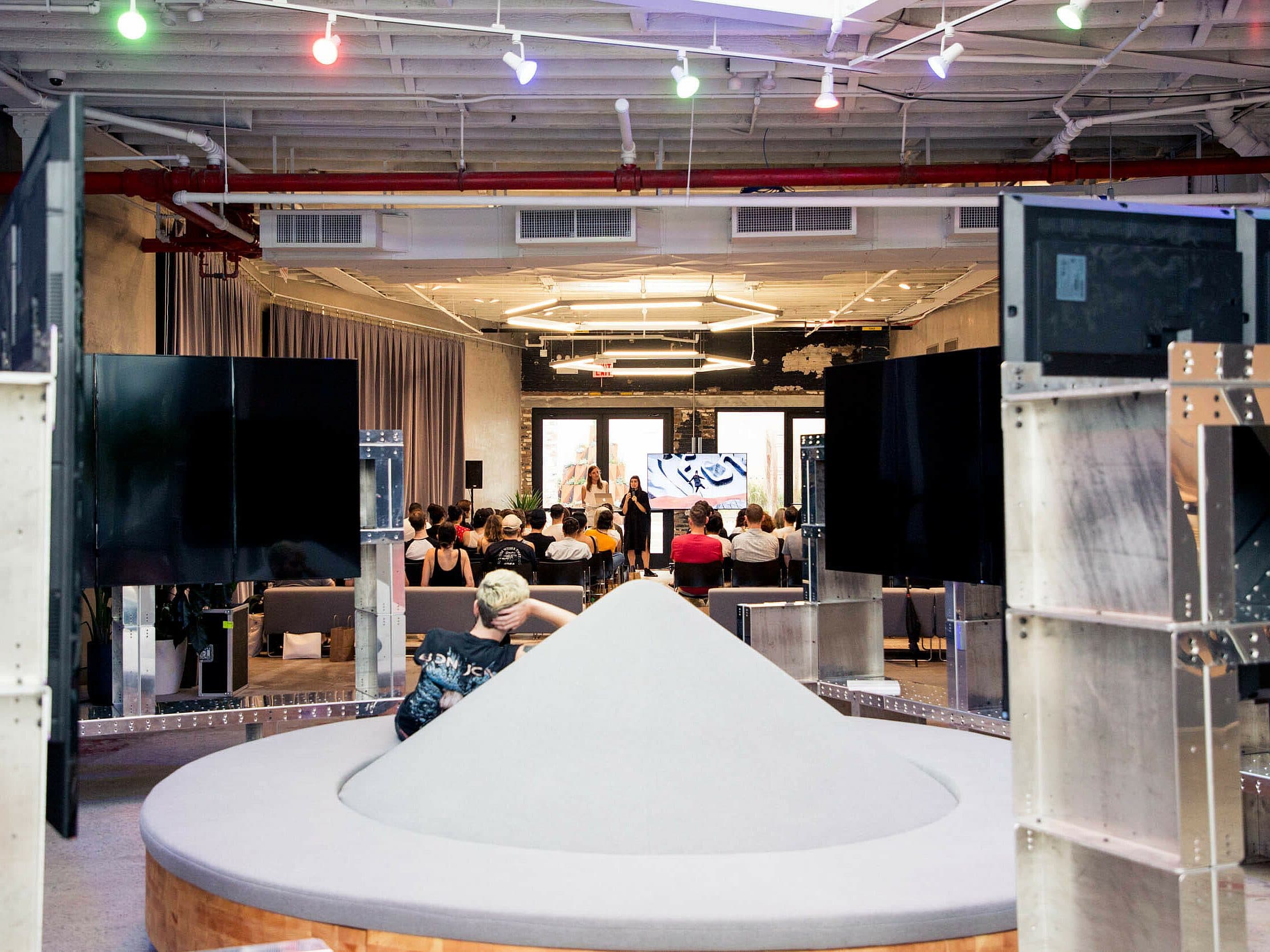 A modern indoor space with an eclectic design features a gathering of people seated and facing a speaker in the background. The room has multiple screens and colorful ceiling lights. In the foreground, there is a large circular structure with a central conical display.