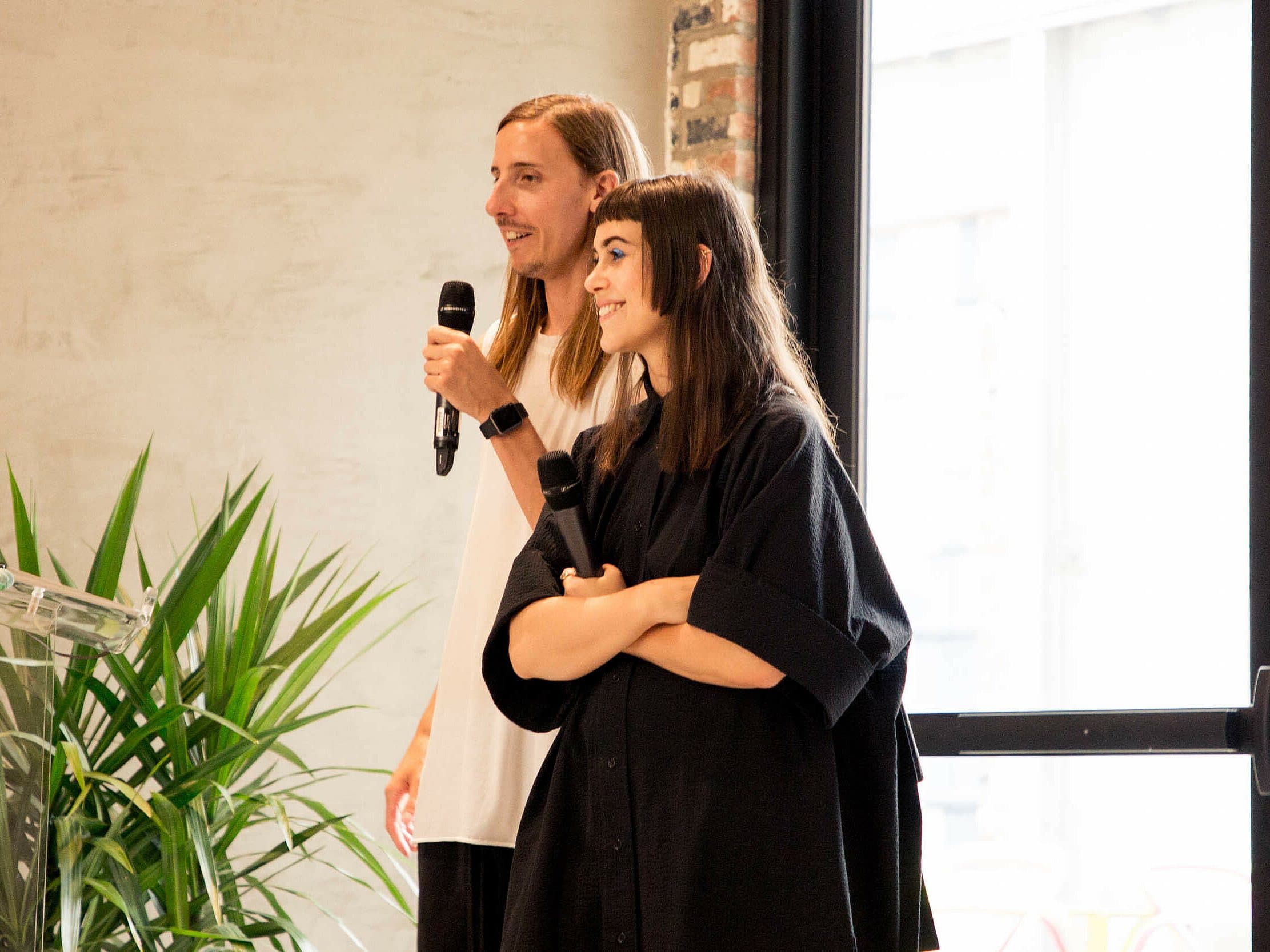 A man and a woman stand side by side holding microphones in a well-lit room. The man has long hair and wears a white shirt, while the woman, with shoulder-length hair, is dressed in black with her arms crossed. A plant and a window are visible in the background.