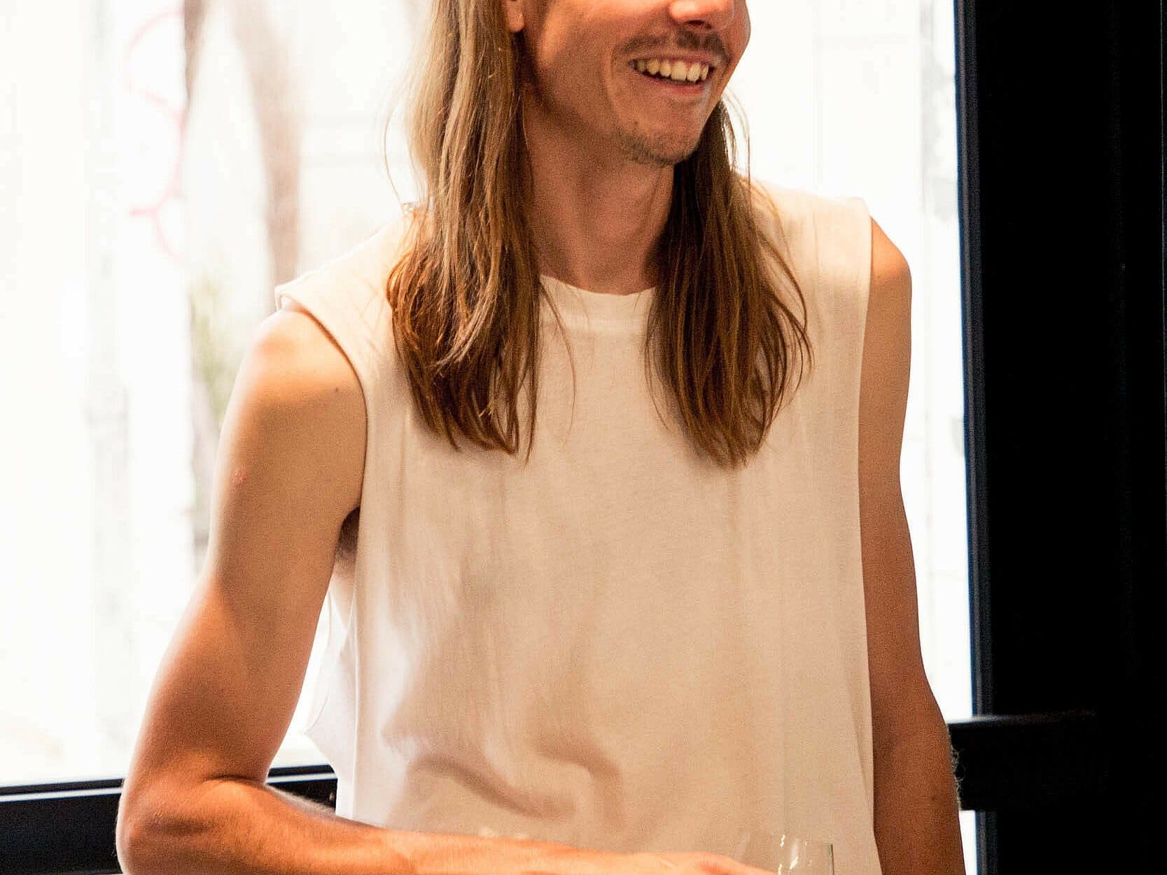 A person with long hair and wearing a sleeveless white shirt is standing indoors, smiling, and holding a glass. The background features a window revealing some outdoor elements in soft focus.