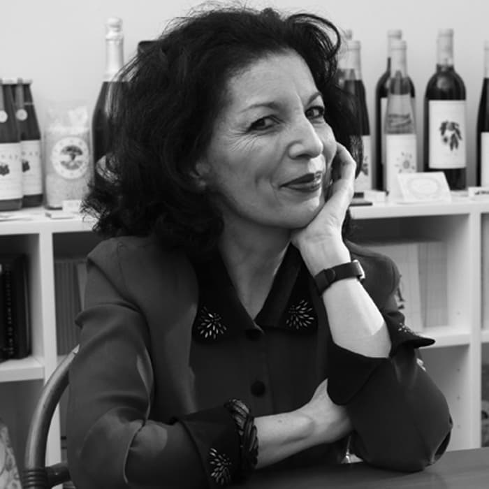 A woman with shoulder-length dark hair is seated, resting her face on her hand, and smiling slightly. She is wearing a dark outfit with decorated buttons. Behind her are shelves with various wine bottles and items arranged neatly. The image is in black and white.