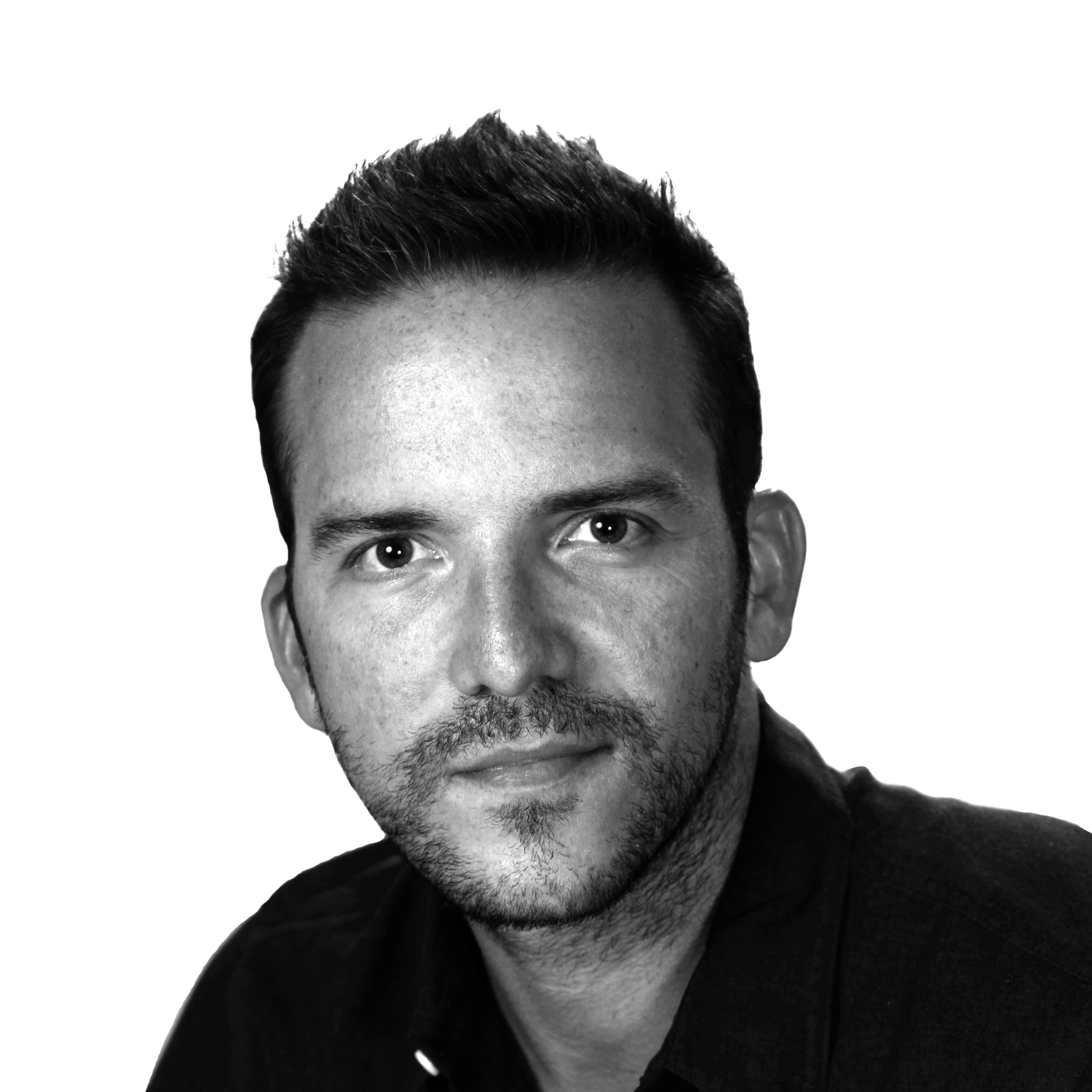 Black and white portrait of a man with short, dark hair and a neatly trimmed beard. He has a calm expression and is looking directly at the camera. He is wearing a dark-colored, button-down shirt. The background is plain and white.