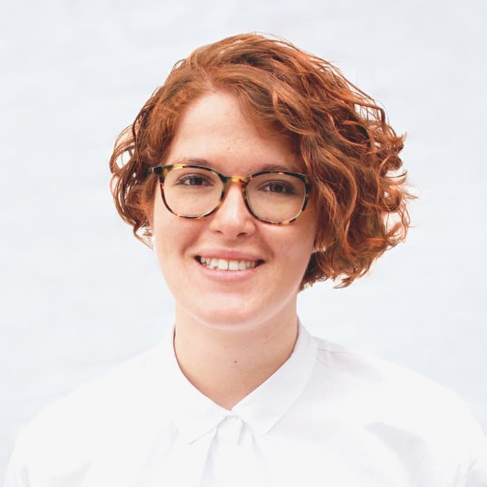 A person with short, curly red hair and wearing glasses is smiling at the camera. They are dressed in a white button-up shirt and are standing against a light-colored background.