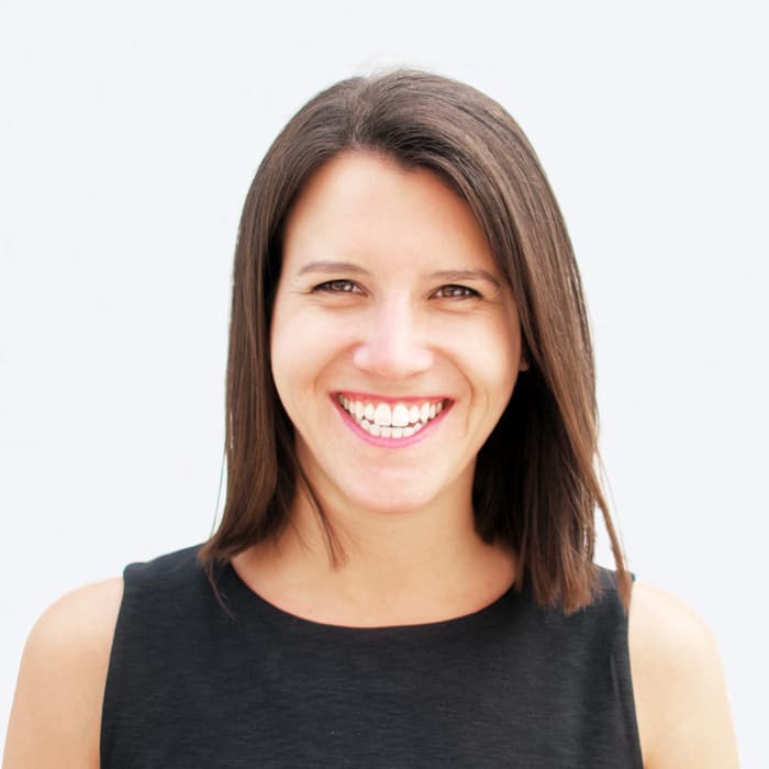 A woman with shoulder-length brown hair is standing against a plain white background. She is wearing a sleeveless black top and smiling brightly at the camera.