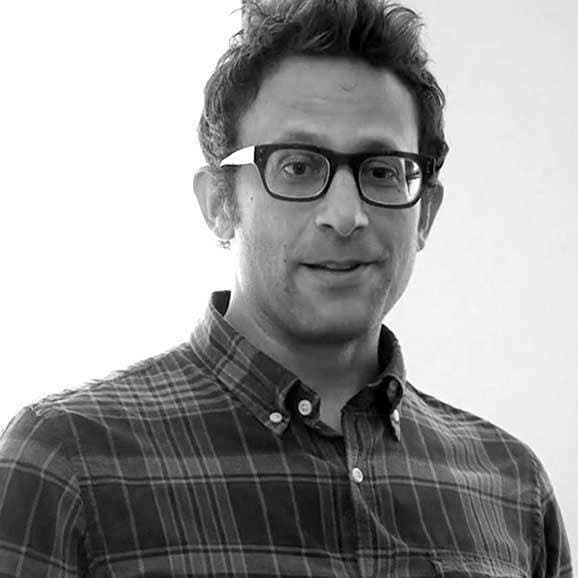 A man with short, tousled hair and glasses is wearing a plaid shirt. He is looking towards the camera with a slight smile. The background is plain and well-lit.