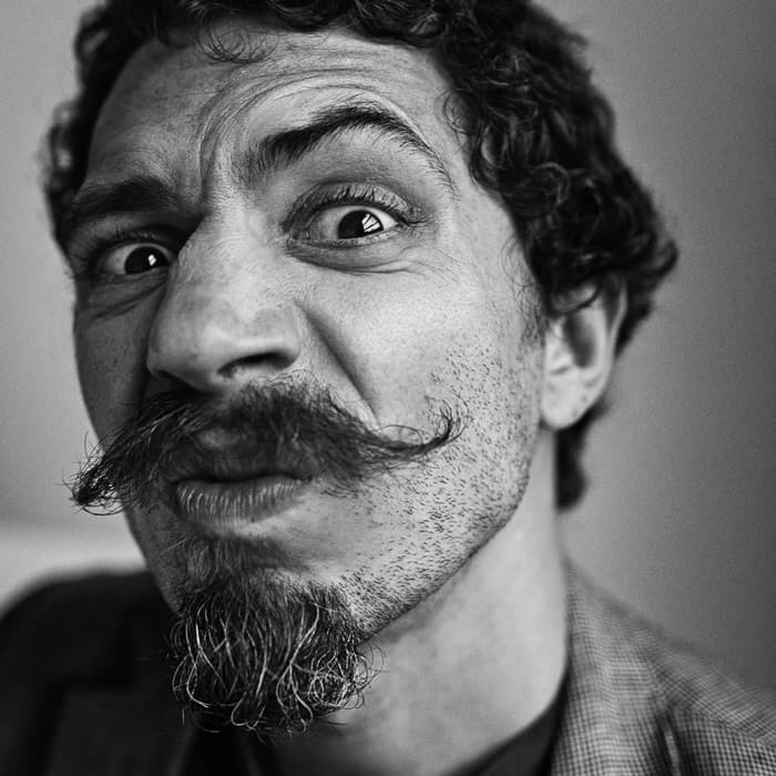 Close-up black and white photo of a man with curly hair and a thick mustache, making a playful, exaggerated facial expression. He has a short beard and is wearing a suit jacket. His eyes are wide open, and he's slightly leaning forward toward the camera.