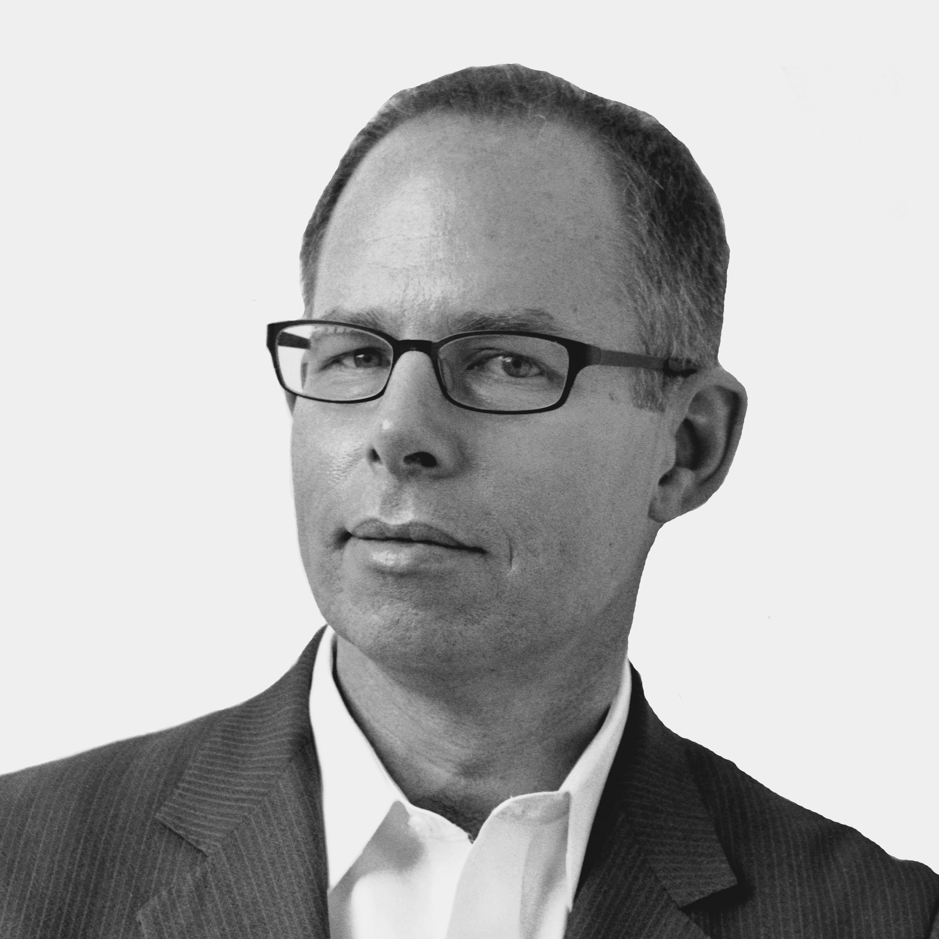 A man with short hair and glasses wearing a suit jacket over a buttoned shirt. He has a neutral expression and is facing slightly to his left against a plain white background.