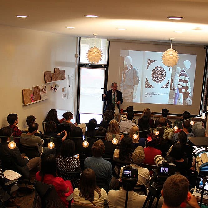 A group of people sit in a cozy, brick-walled room, attentively listening to a speaker standing in front of a projection screen. The screen displays an image and text. The room is decorated with string lights and has a warm, intimate atmosphere.