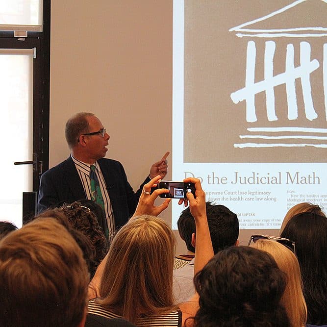 A man in a suit and tie stands at the front of a room, giving a presentation to a seated audience. He gestures towards a projected slide on the wall behind him, which features text and an illustration resembling a courthouse. Audience members hold up cameras.