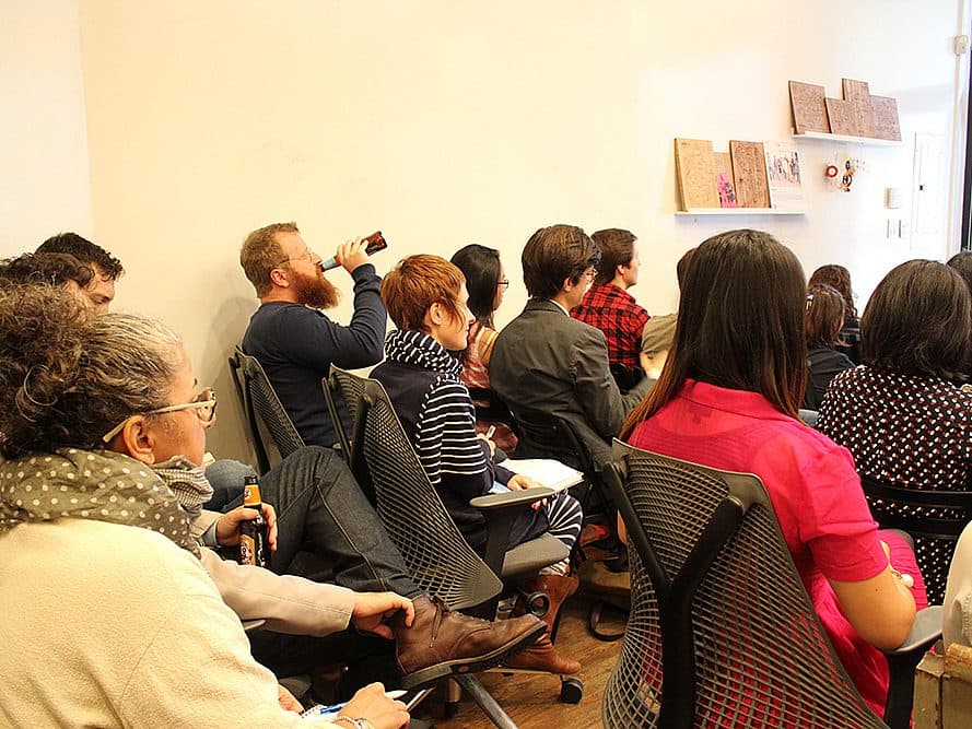 A group of people sitting closely together in a small room, attentively watching a presentation. Some are taking notes while others are holding drinks. The setting is informal, with casual attire and simple decor on the walls.
