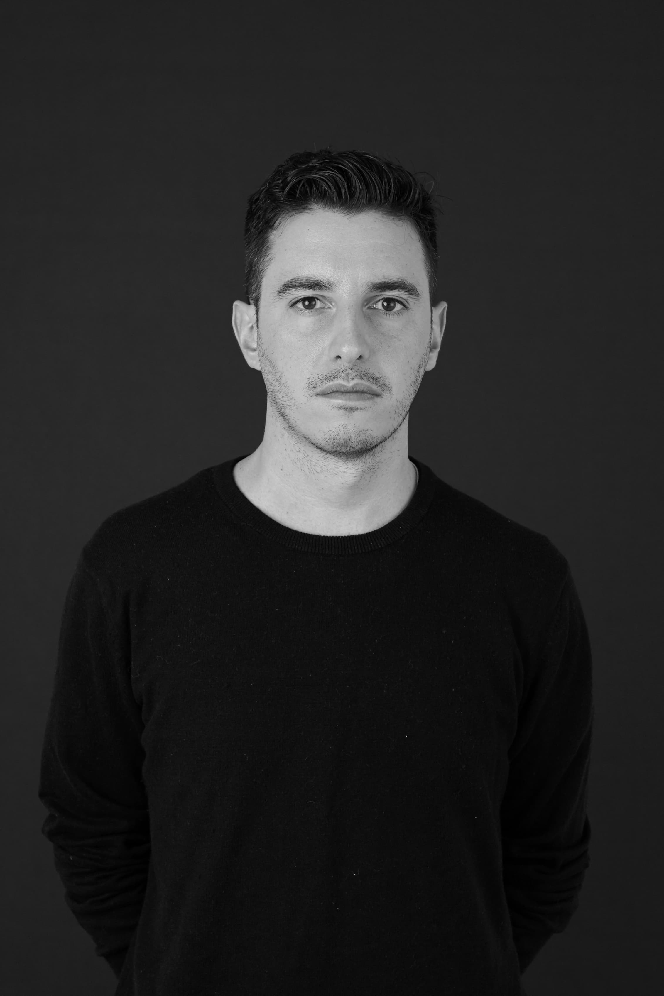 A black-and-white portrait shows a man with short, dark hair and facial stubble. He is wearing a plain, dark-colored sweater. The background is solid black. The man looks directly at the camera with a neutral expression, his arms relaxed at his sides.