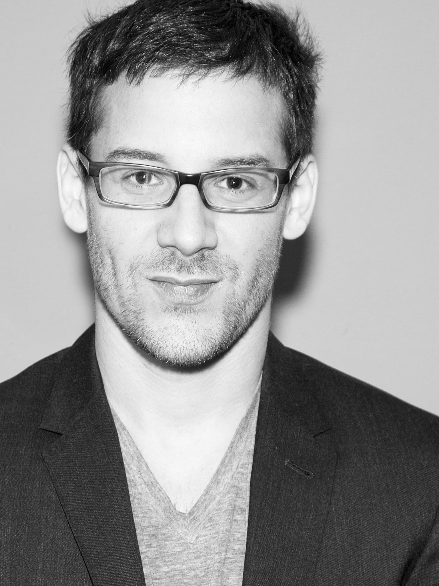 A black-and-white photo of a man with short hair and glasses. He is wearing a dark blazer over a V-neck shirt. The man is looking directly at the camera with a slight smile. The background is plain and light-colored.