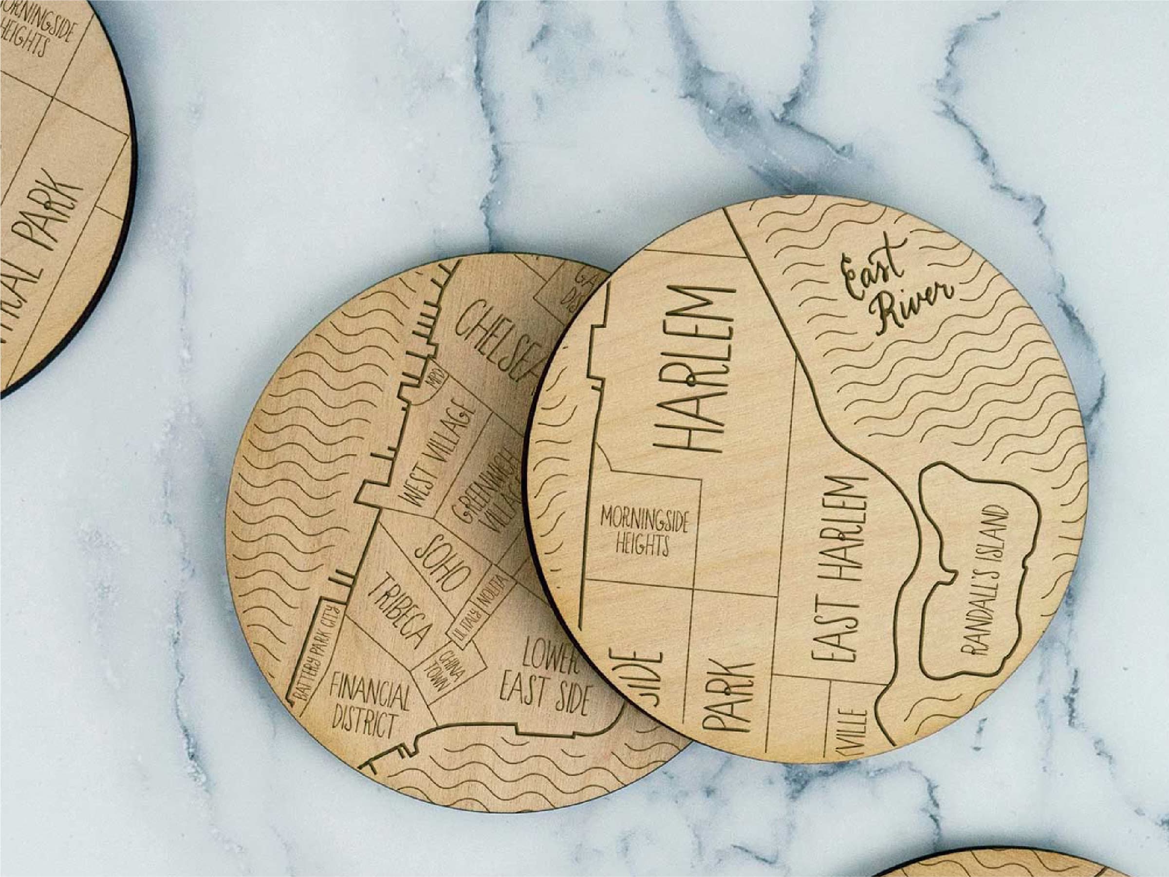 A set of round wooden coasters engraved with a map of New York City's boroughs and neighborhoods, including Harlem, East Harlem, Chelsea, Tribeca, and Soho. The coasters rest on a white marble surface.