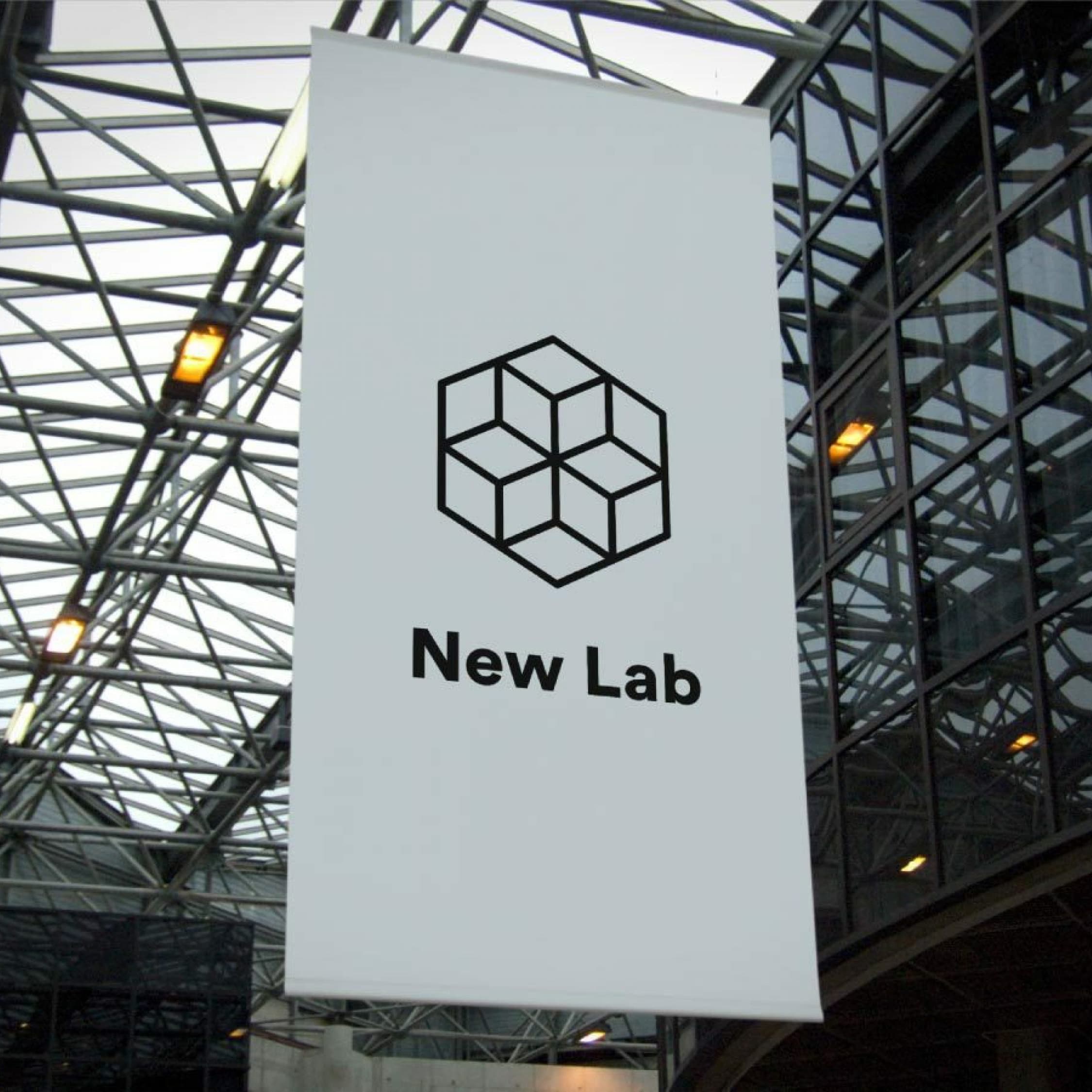 A vertical banner hanging in an indoor atrium with a glass and metal roof. The banner features a geometric cube logo above the text "New Lab" in black. The atrium structure is contemporary, with glass panels and metal beams.