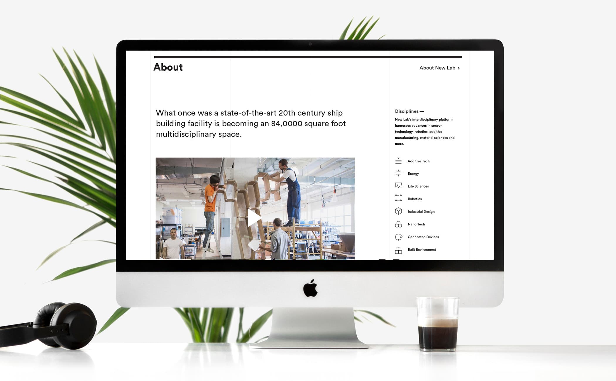 A computer monitor displays a webpage with a section titled "About." The text describes a former 20th-century shipbuilding facility being converted into an 84,000-square-foot multidisciplinary space. A photo shows people working on a large sculpture. Headphones and a glass are beside the monitor.