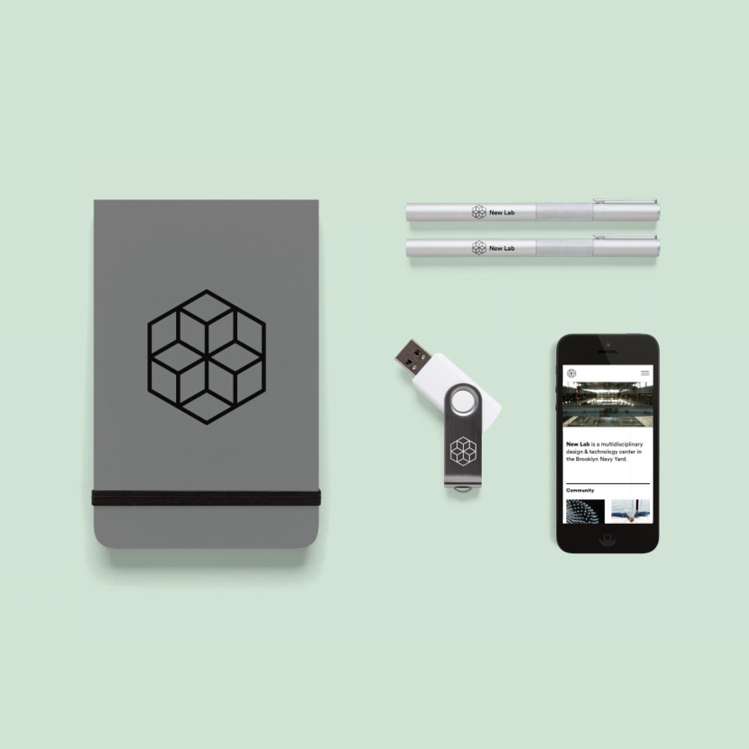 A flat lay of branded stationery including a gray notebook with a black geometric logo, two white pens, a USB flash drive, and a smartphone displaying a website on a mint green background.