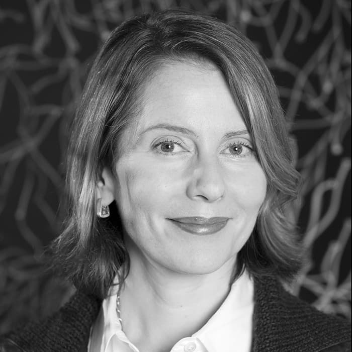 A black and white portrait of a person with a slight smile. They have shoulder-length hair and are wearing a white collared shirt with a dark sweater. The background features an abstract, twisting line pattern.