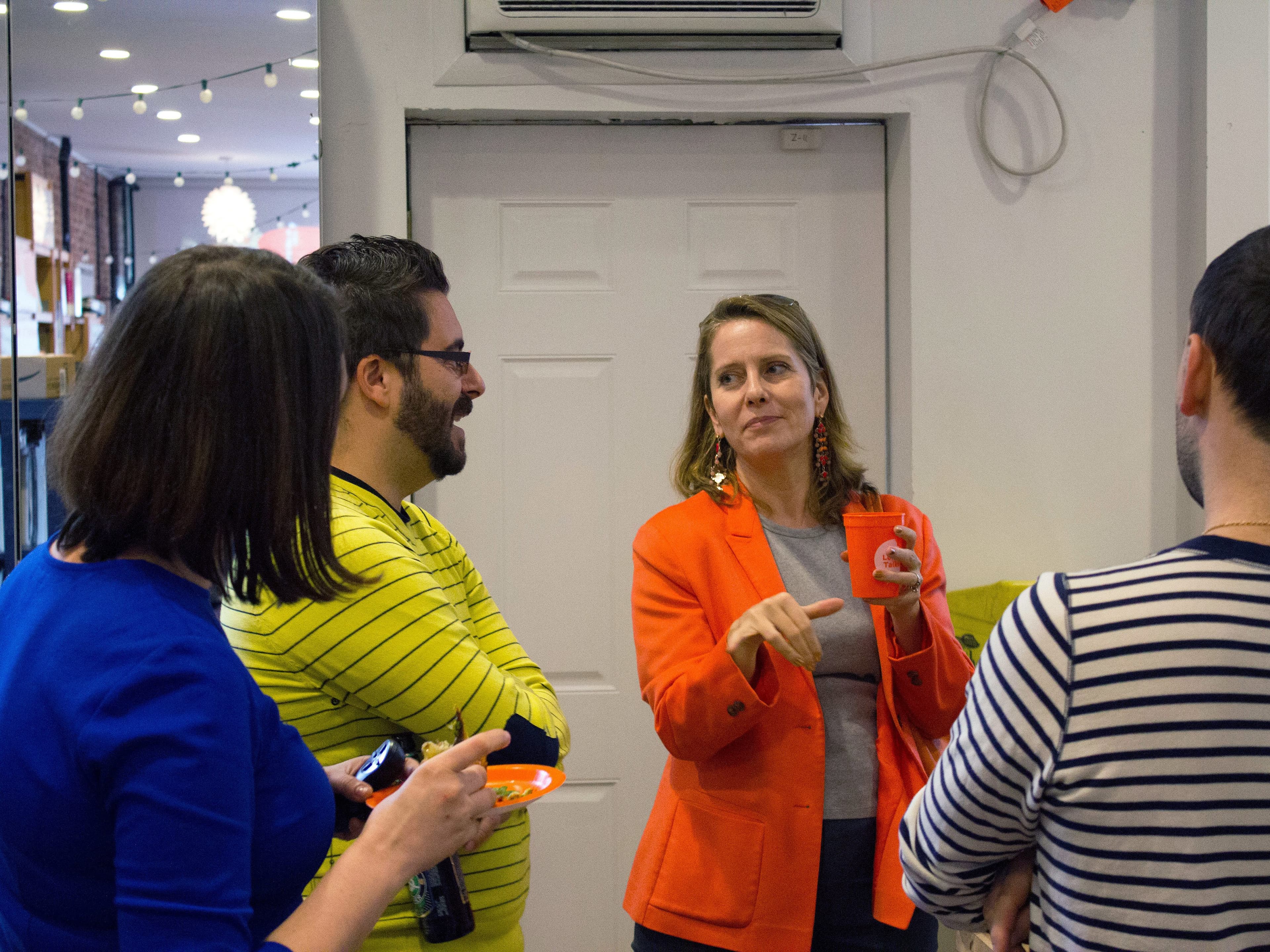 A group of four people engaged in a conversation indoors. One woman, wearing an orange blazer and holding a red cup, appears to be speaking, while others listen attentively. The room is well-lit with shelves and decorations visible in the background.