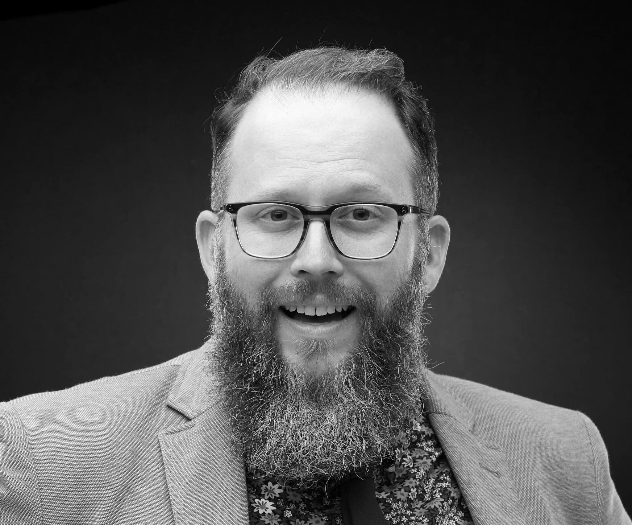 A black and white portrait of a person with short hair wearing glasses and a full beard. The individual is dressed in a blazer and a patterned shirt, smiling with an open mouth against a dark background.