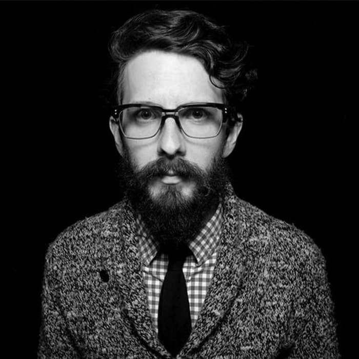 Black-and-white portrait of a man with wavy hair and a full beard wearing glasses. He is dressed in a checkered shirt, a dark tie, and a textured blazer. The background is black, drawing focus to the individual.