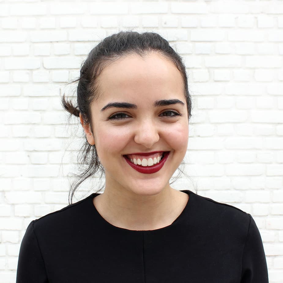 A young woman with dark hair pulled back is smiling warmly at the camera. She is wearing a black top and has red lipstick. The background is a white brick wall.