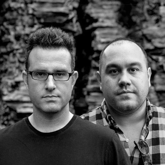 Two men stand outside against a backdrop of stacked rocks. The man in the foreground has short hair, glasses, and is wearing a dark T-shirt. Behind him, the second man has a shaved head and wears a checkered shirt with sunglasses hanging from the collar. Both look serious.