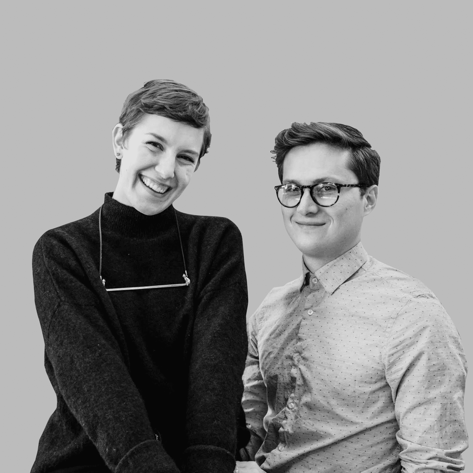 Black and white image depicting two smiling individuals. The person on the left has short hair and wears a dark sweater with a string of glasses around their neck, sitting with their hands clasped together. The person on the right wears glasses and a light polka-dotted shirt.