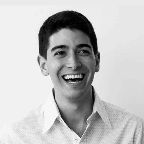 Black and white photo of a young man with short dark hair, smiling broadly and looking off to the side. He is wearing a light-colored, button-up shirt with a subtle pattern. The background is plain and white.
