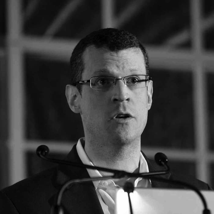 Black and white photo of a man speaking into a microphone. He is wearing glasses, a suit, and a white shirt. The background is blurred, with what appears to be window panes allowing light to filter through. The image conveys a professional or formal setting.