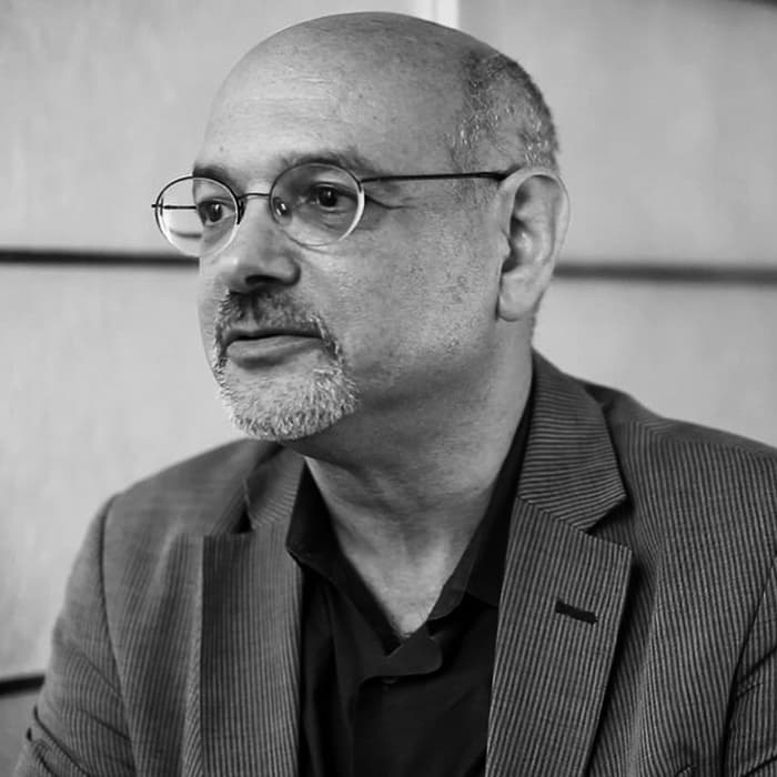 A black and white image shows a man with glasses, a short beard, and a bald head. He is wearing a striped blazer and a dark shirt. The person looks to the side with a calm and contemplative expression. The background is blurred, focusing attention on the individual.