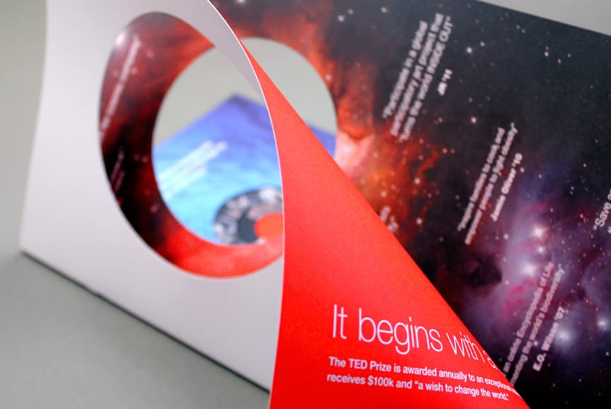 A close-up shot of a uniquely designed flat envelope with round cut-outs and cosmic-themed print. The open red flap reveals text about the TED Prize, including the award amount of $100K and the transformative goal of the recipient's wish.