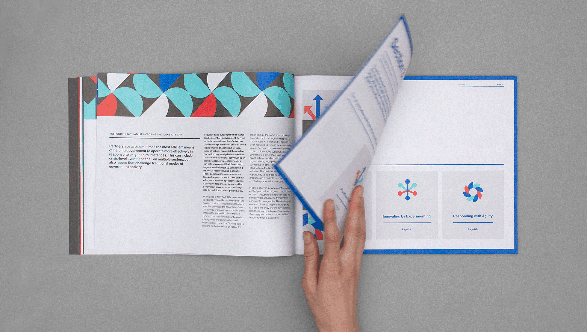 A person's hand is seen flipping the page of an open book or magazine. The left page displays text and a geometric, colorful pattern. The right page features diagrams with colorful circular designs. The background is plain and light gray.