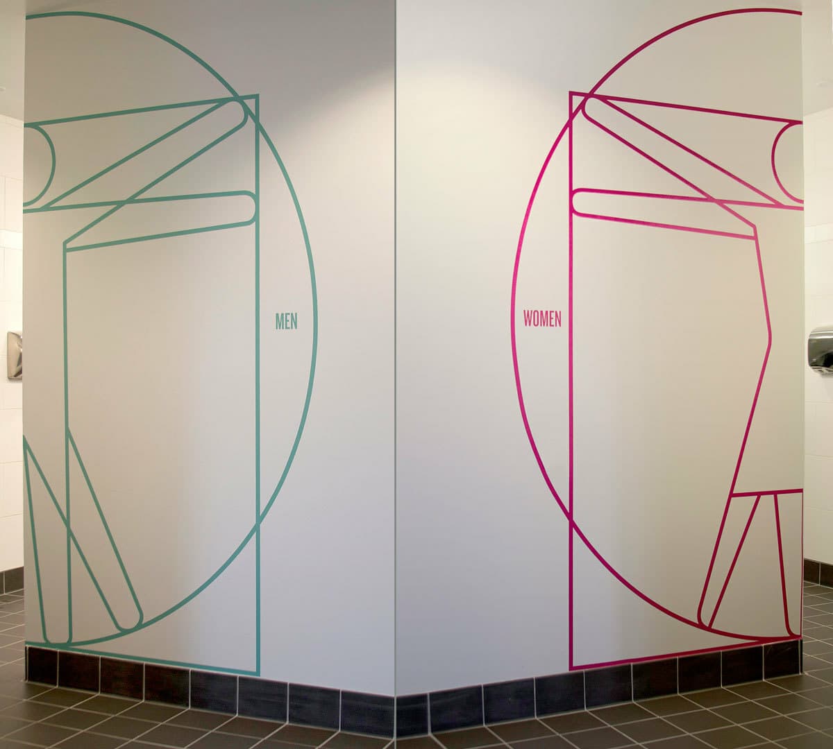 A restroom entrance shows modern, artistic representations of men and women. The men's graphic is in teal, and the women's graphic is in magenta. Each design uses geometric shapes to form a human figure and is labeled accordingly.