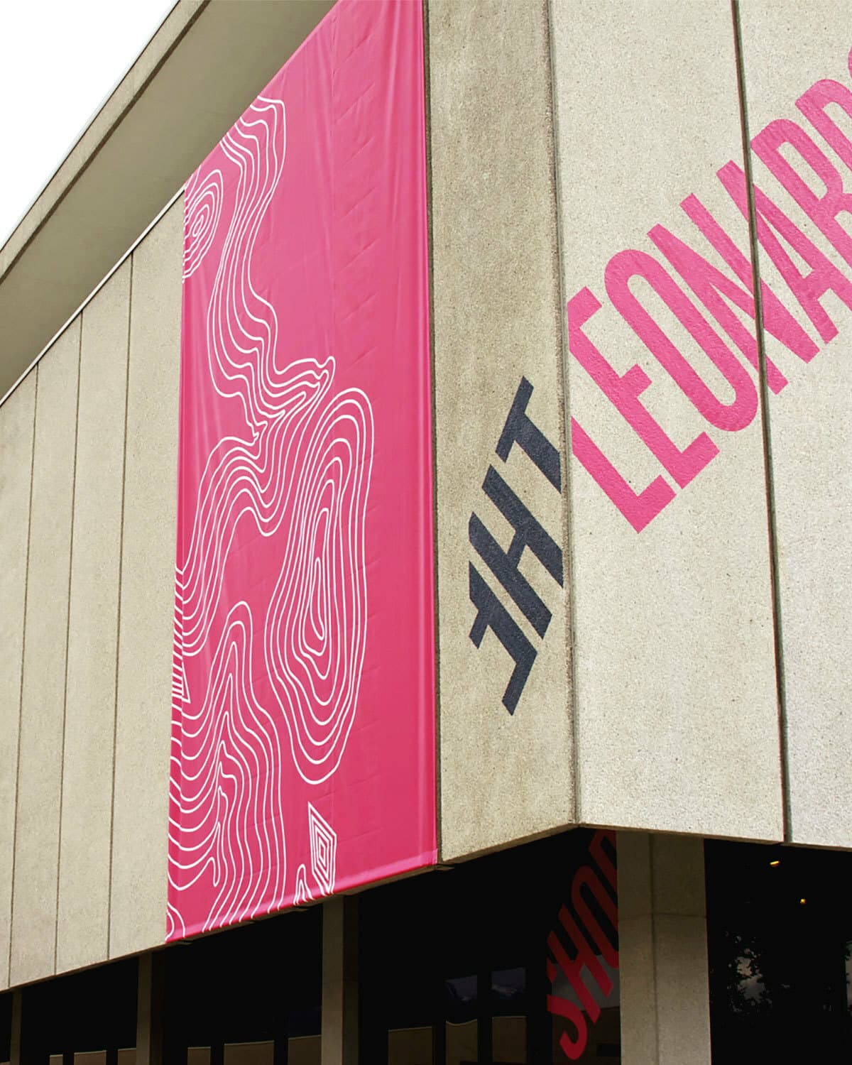 A beige concrete building with a large vertical pink banner displaying abstract white line art. The corner of the building has the word "LEONARD" in large pink letters and "THE" in smaller black letters. A storefront with glass windows is visible below.