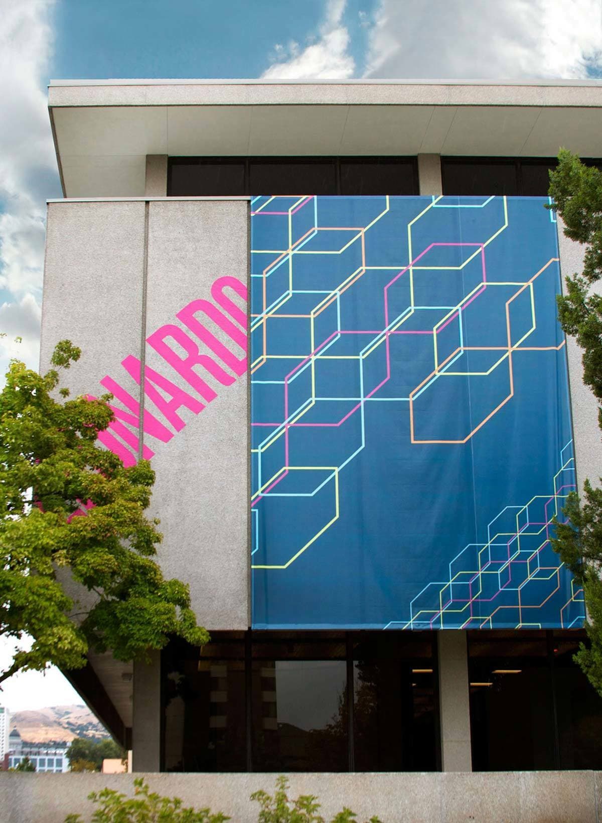 A large concrete building with an artistic banner displayed on its side. The banner features a geometric design with interconnected cubes in various colors against a blue background. The word "LEONARDO" is vertically written in large, bold pink letters on the left.