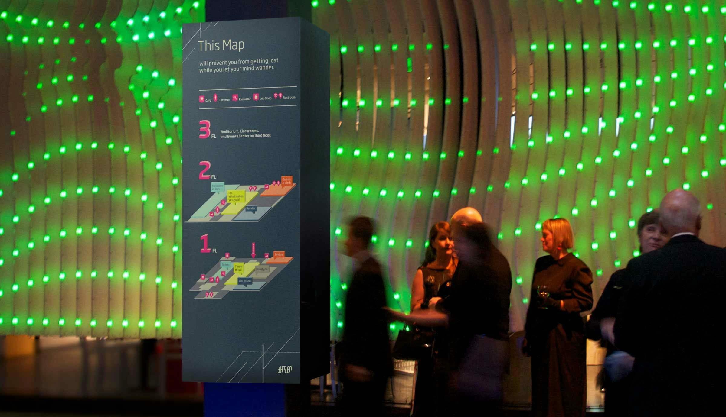 A large information stand displays a map of an indoor location with three numbered steps. The background features a wall adorned with green illuminated dots. Several people, some in motion and others standing, are gathered around the sign.