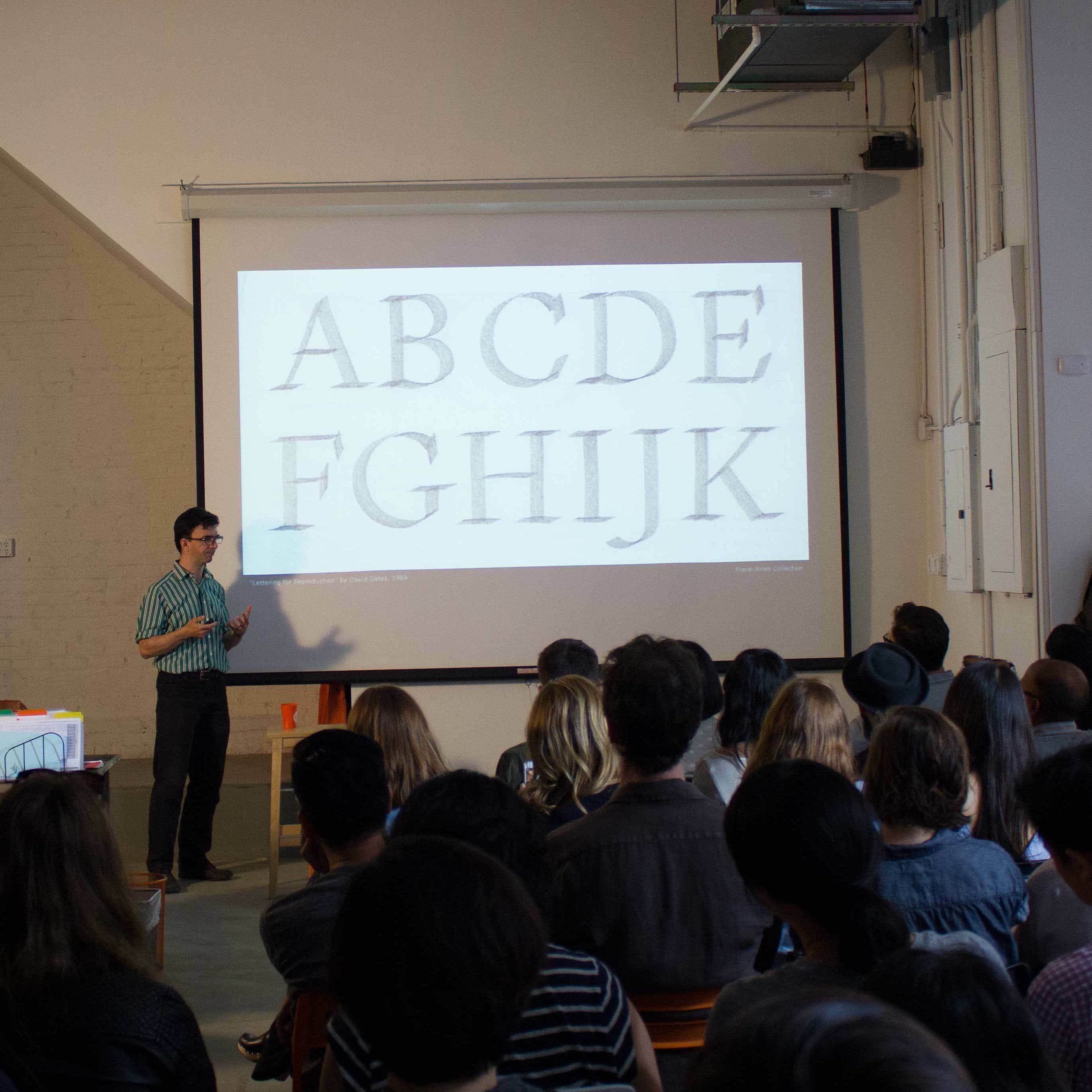 A person stands in front of an audience giving a presentation. On the projector screen behind them is a slide displaying large uppercase letters from A to K. The room is filled with seated attendees, attentively listening to the speaker.