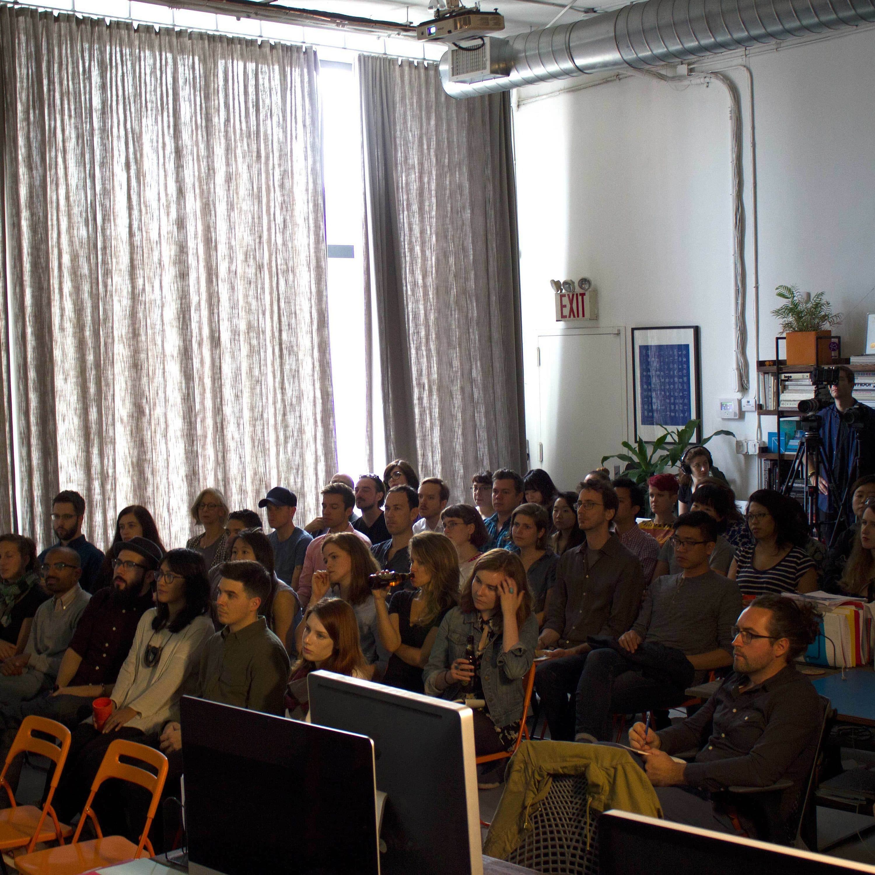 A group of people, sitting closely together on chairs, attentively listen to a speaker in a dimly lit room with large windows covered by curtains. The room has a casual setup with desks and computer monitors in the foreground and a camera on a tripod at the back.