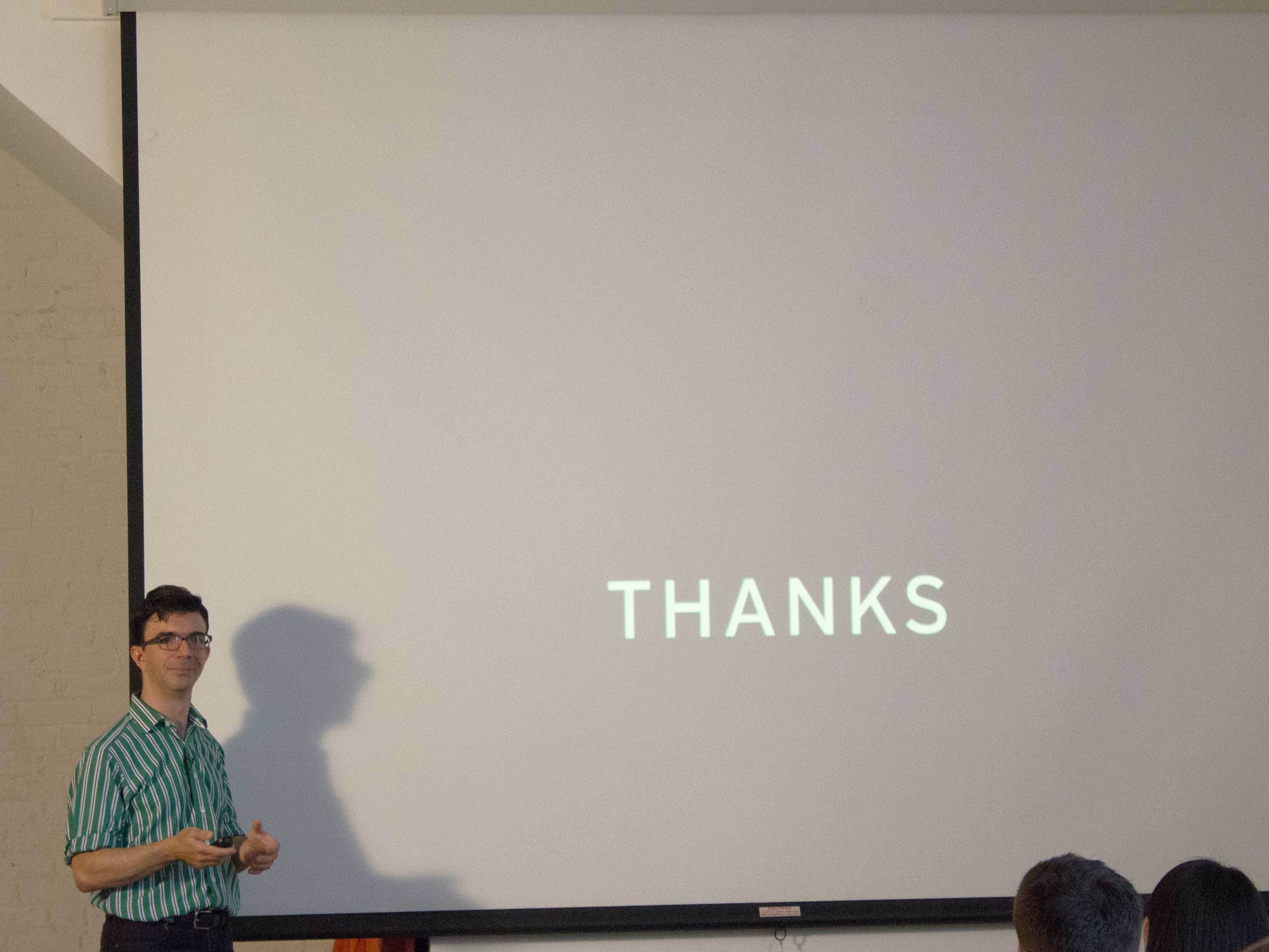 A person in a green and white striped shirt stands by a large screen during a presentation. The screen displays the word "THANKS" in capital letters. An audience member's head is partially visible in the foreground.