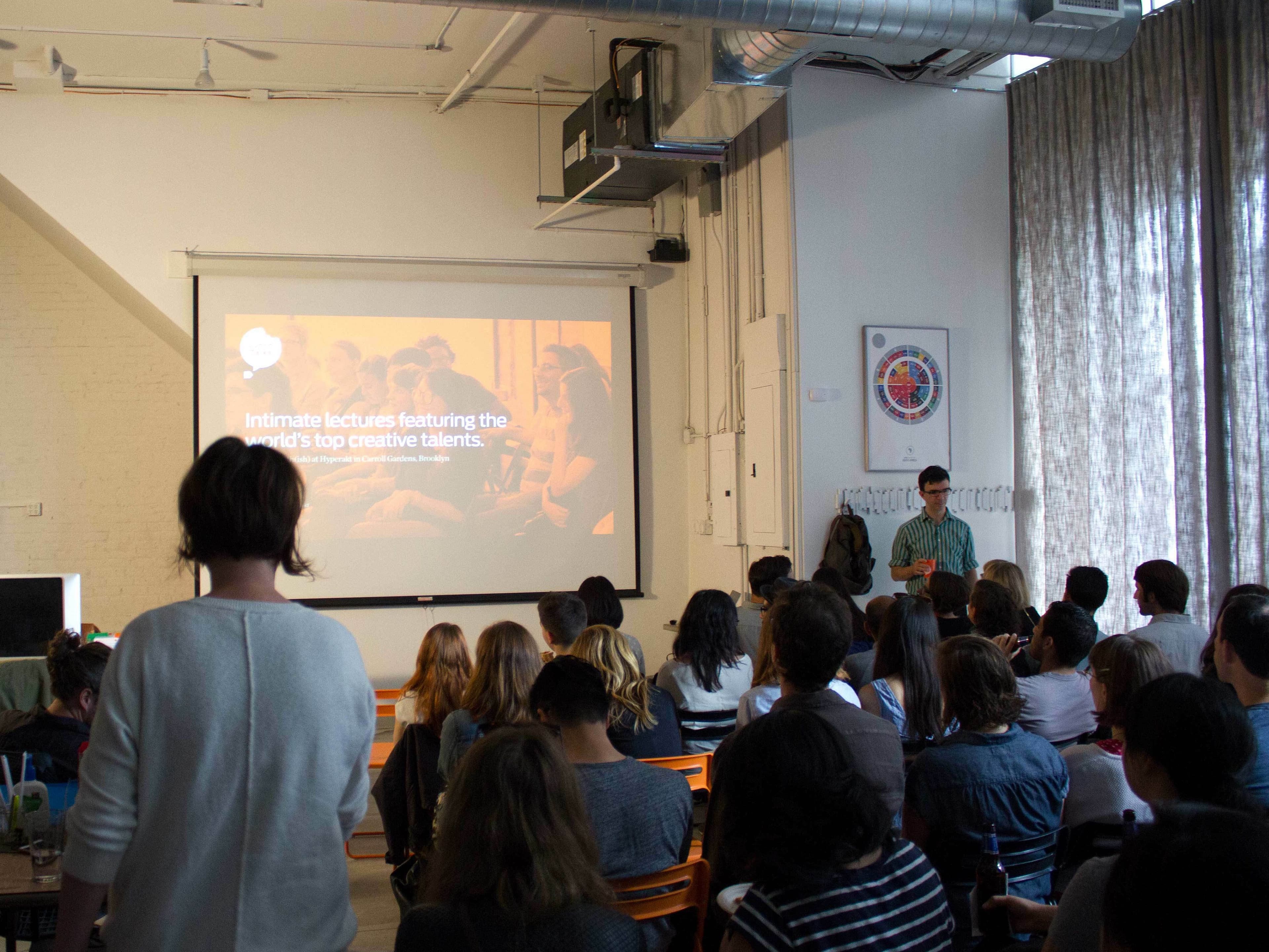 A group of people sitting in rows of chairs attentively listen to a presenter standing near a projection screen. The screen displays a slide with text that reads, "Intimate lectures featuring the world’s top creative talents." The room has high ceilings and large windows.