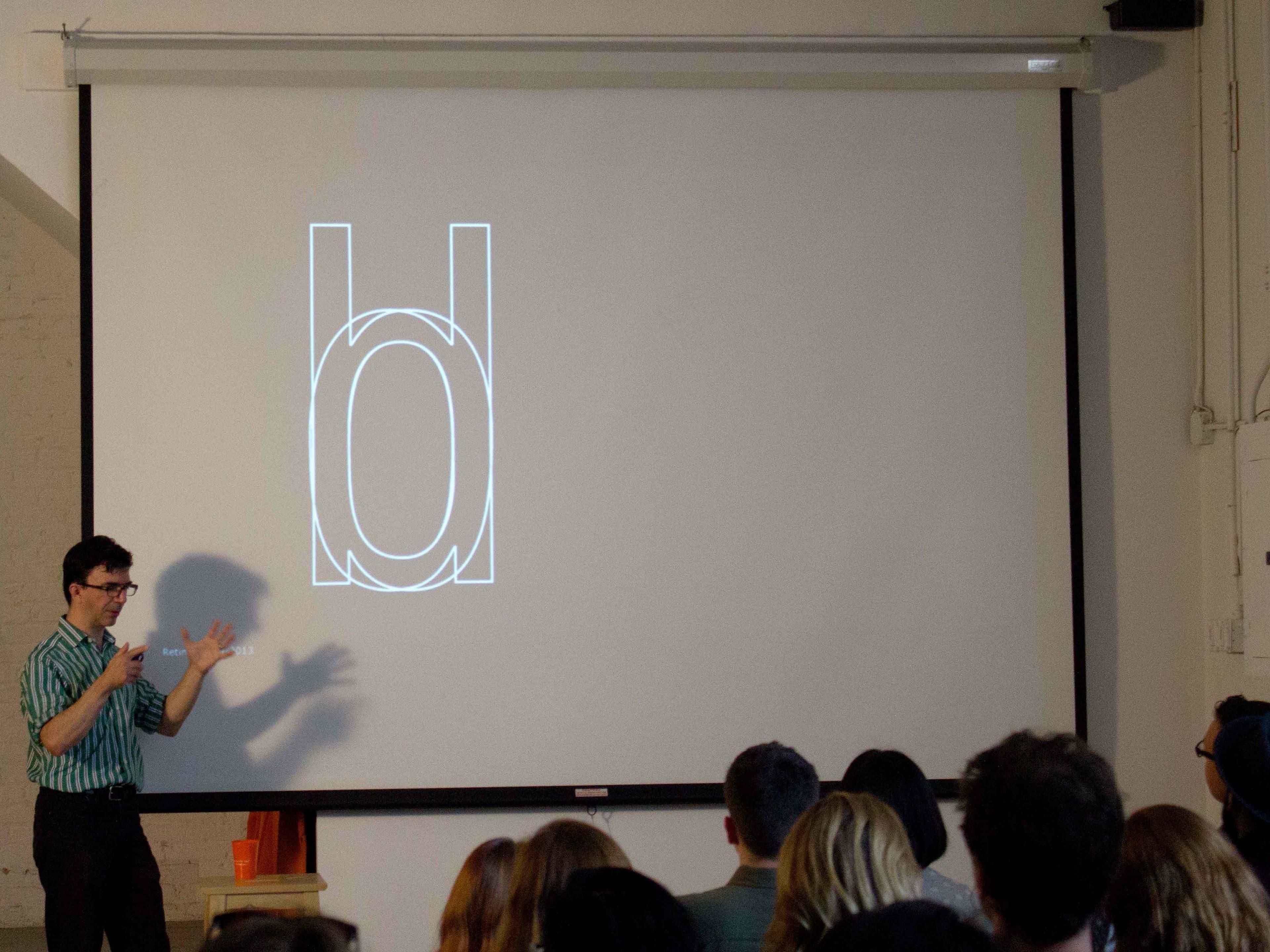 A person wearing a striped shirt is presenting in front of an audience. They are gesturing with their hands towards a large screen displaying a stylized letter "d" with overlapping geometric shapes. The audience is seated and observing the presentation attentively.