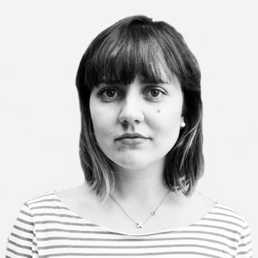 A black-and-white portrait of a person with shoulder-length hair and bangs. They are wearing a striped top and a simple necklace. The background is plain white. The person has a neutral expression.