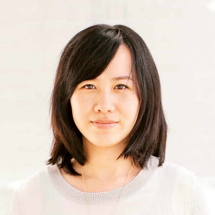 A woman with shoulder-length black hair and bangs, wearing a white top, stands against a light-colored, blurred background. Her expression is neutral, and she looks directly at the camera.