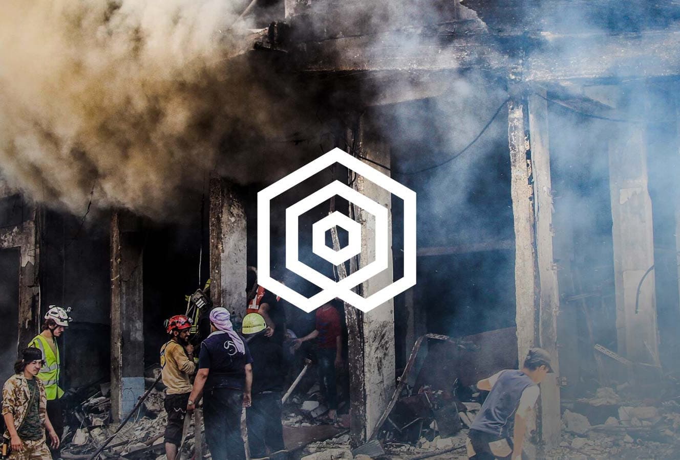 A group of people, some in safety gear, gather amidst debris and smoke in a damaged building. Thick smoke partially obscures the scene. At the center of the image, there is a superimposed geometric logo consisting of concentric hexagons.