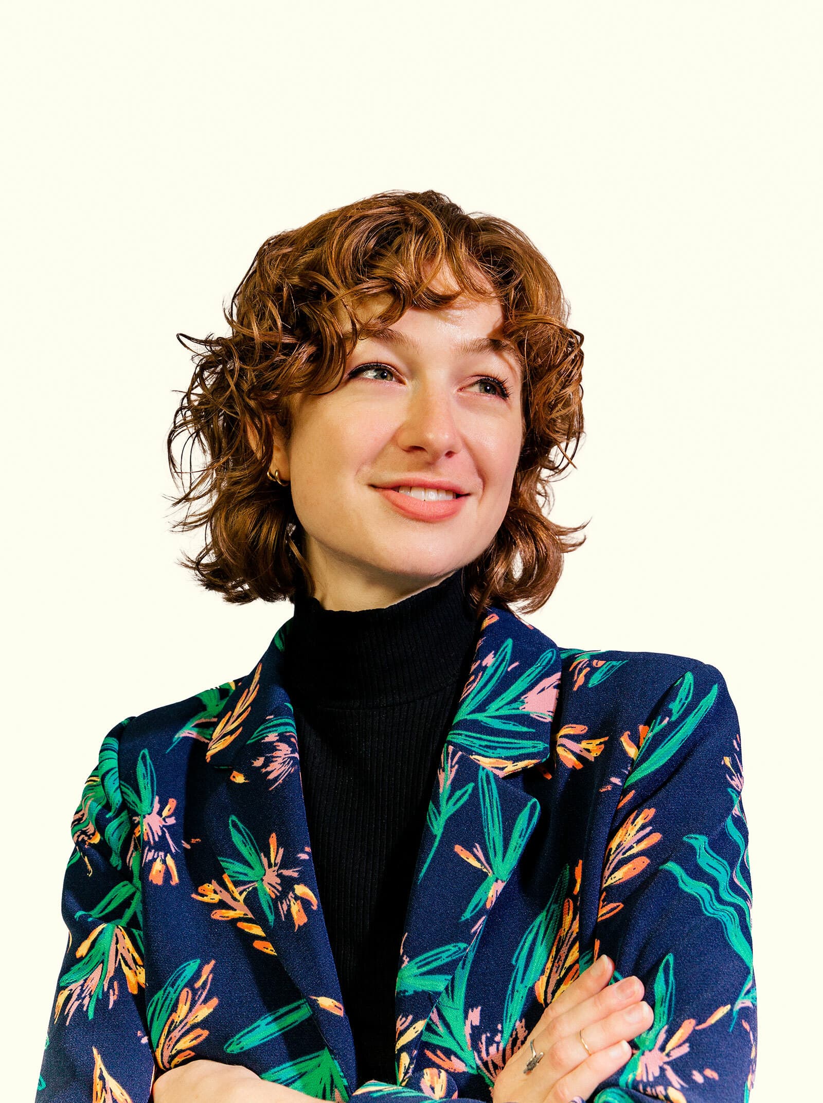 A person with wavy light brown hair wearing a black turtleneck and a colorful floral patterned jacket, smiles and looks off to the side. They have their arms crossed casually. The background is plain and light-colored.