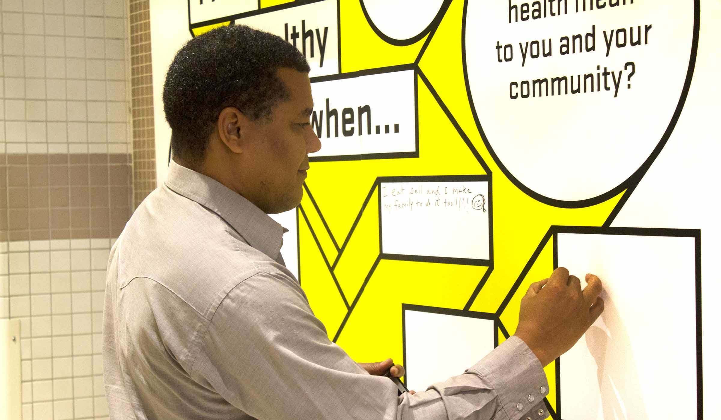 A man wearing a light grey shirt writes on a large, interactive wall display with a speech bubble design. The display features questions about health, prompting participants to fill in their responses.